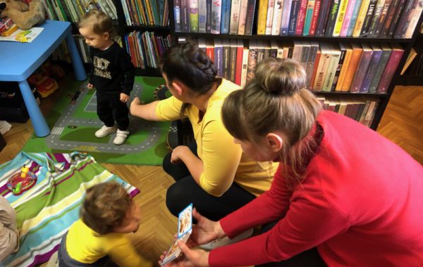 Bibliotekarz pokazujący maluszkowi przygotowaną dla dzieci książeczkę. Z tyłu – mama z dzieckiem