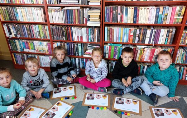 Sześcioro dzieci na dywanie i prezentujących narysowane przez siebie prace. W tle regały z książkami