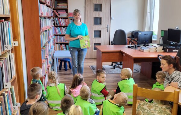 Grupa dzieci sziedząca na podłodze. W tle bibliotekarka prezentująca książkę.