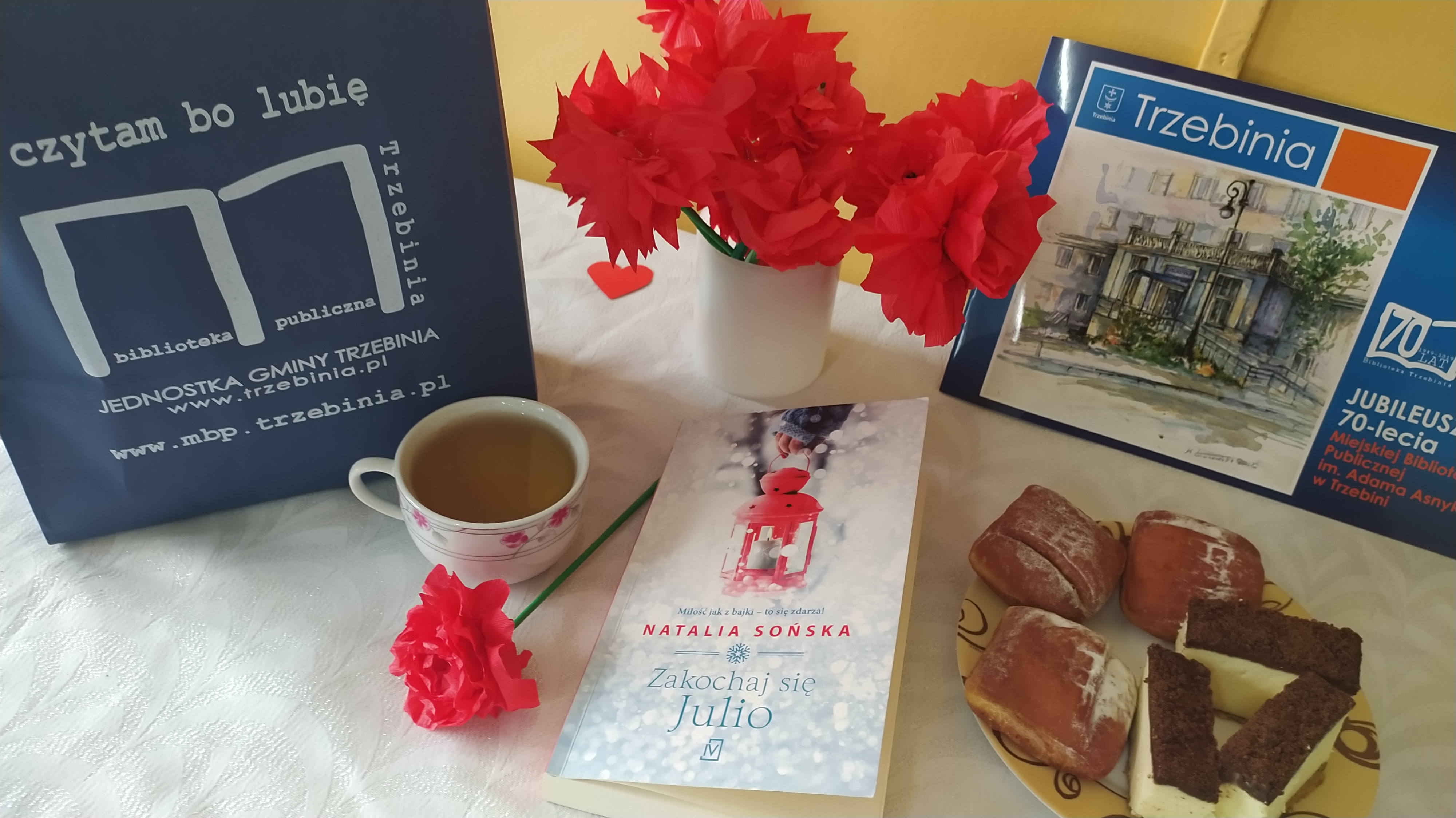 Zdjęcie z niespodzianką przygotowaną dla Jubilatki. Stolik, a na nim książka, filiżanka z herbatą, talerzyk z poczęstunkiem i ozdobne kwiaty z bibuły